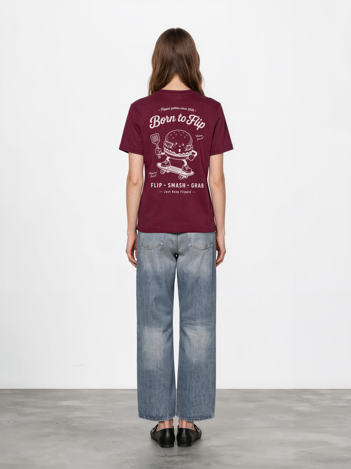 Female model wearing the Born to Flip burgundy T-shirt, back view with large white skateboarding burger artwork across the back.