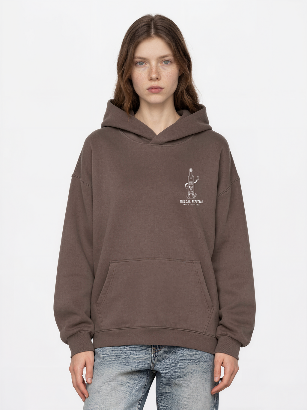 Female model wearing the mocha Holy Smokes oversized hoodie, showing the small white mezcal bottle screen print on the left chest