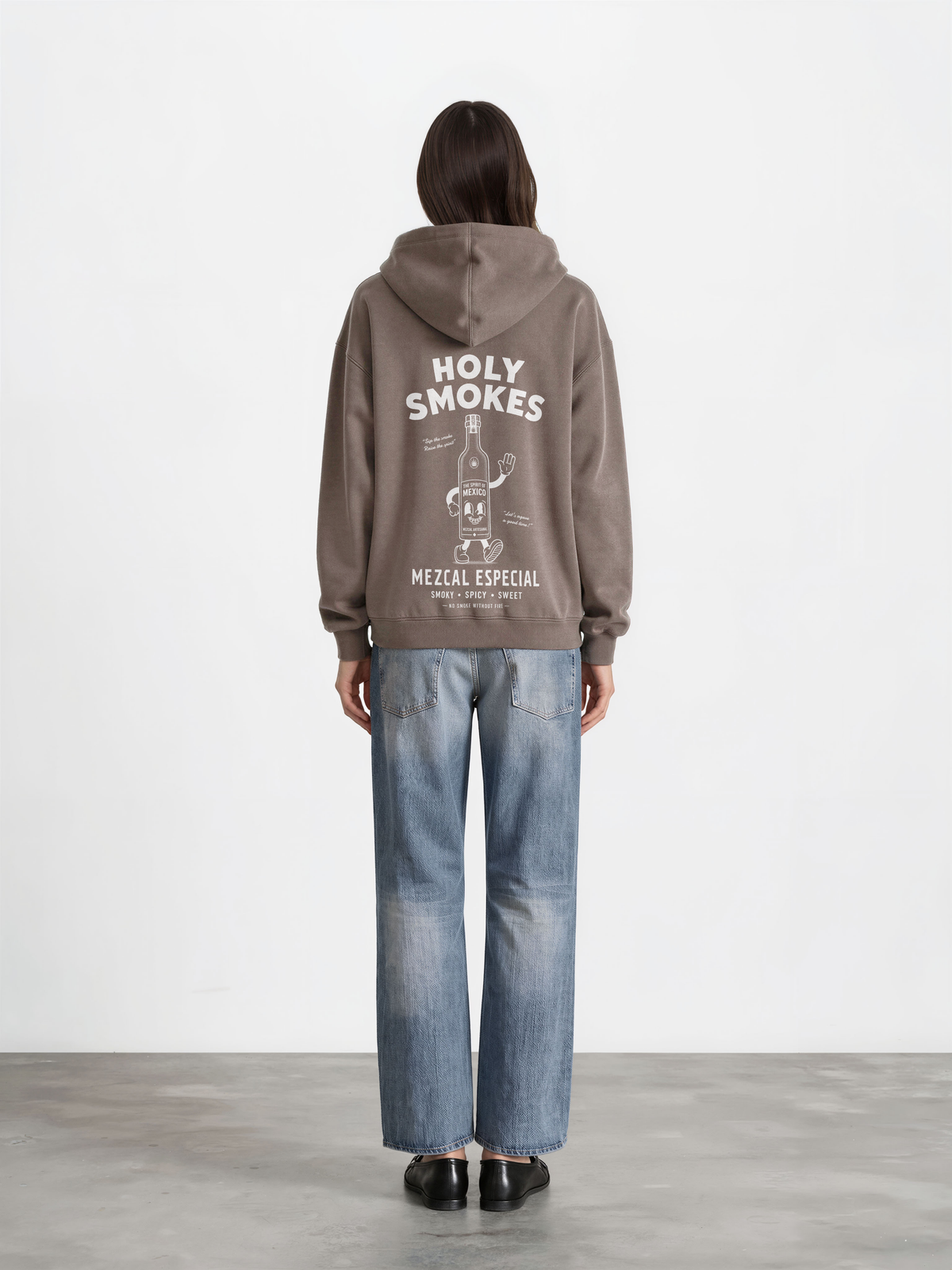 Female model wearing the taupe Holy Smokes oversized hoodie, showing the large white Holy Smokes mezcal bottle screen print on the back