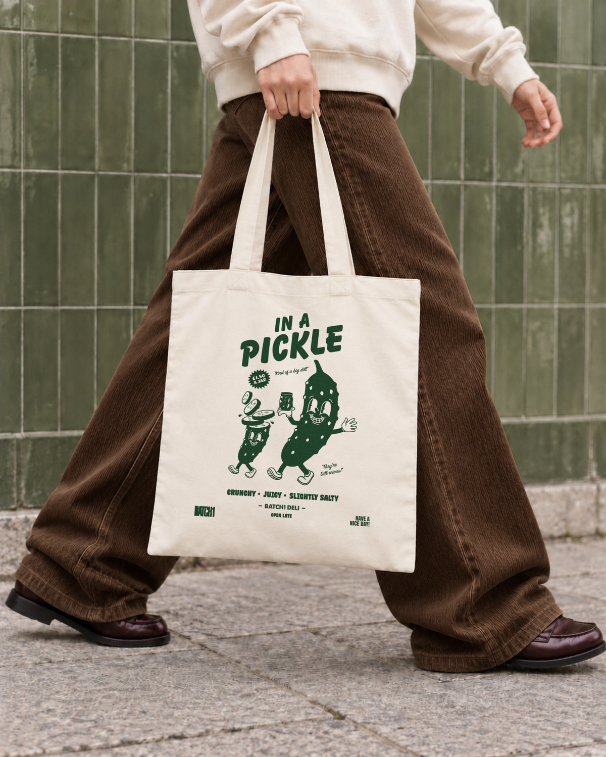 In a Pickle natural cotton tote bag with green pickle artwork carried with chocolate cord trousers and red-brown loafers