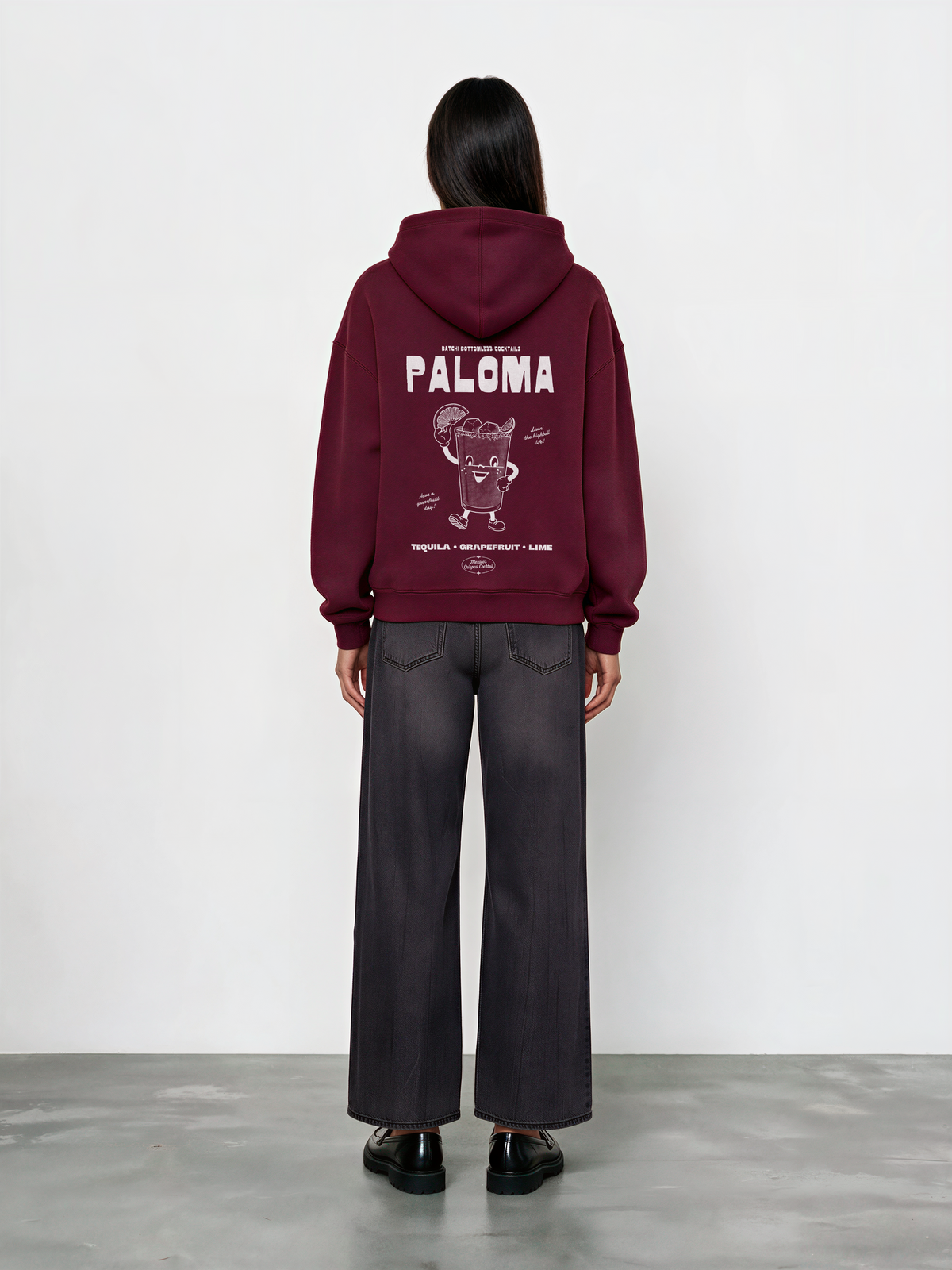 Female model wearing the Paloma burgundy oversized hoodie, back view showing the white cocktail graphic just beneath the hood.