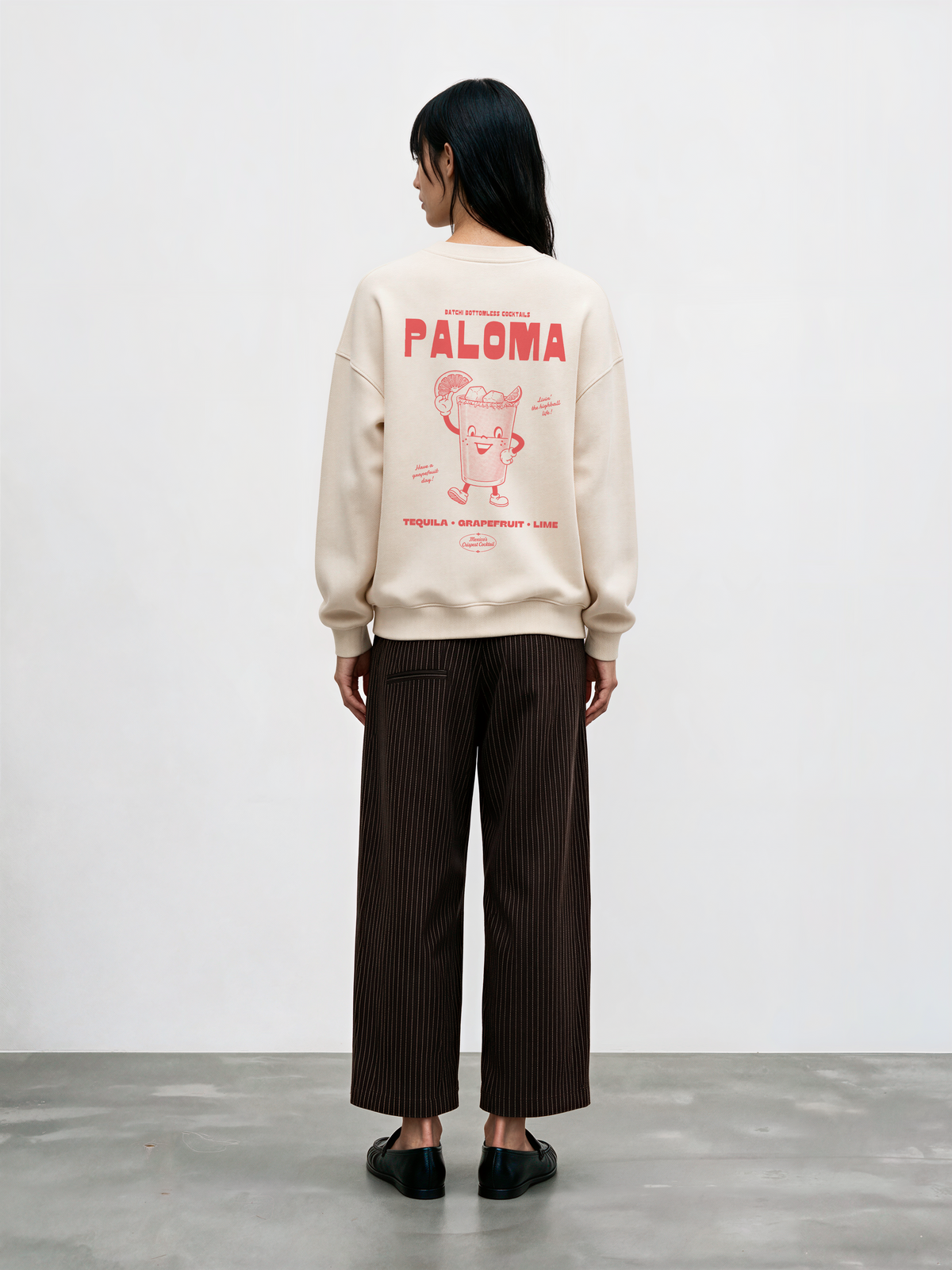 Female model wearing the Paloma ivory oversized sweatshirt, back view showing the coral Paloma cocktail character and tequila, grapefruit and lime lettering.