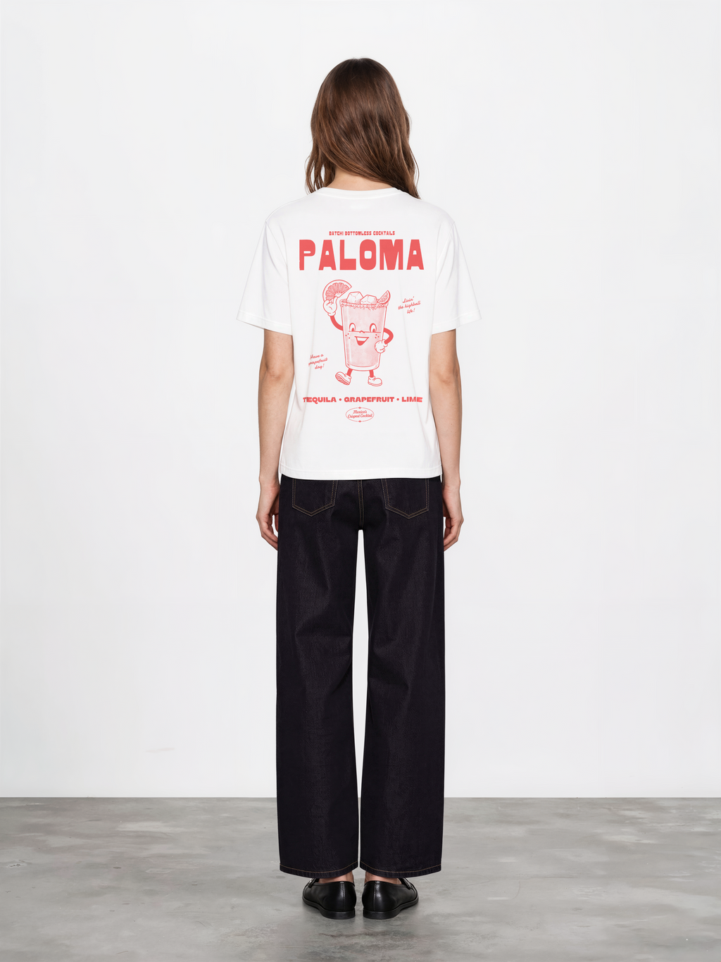 Female model wearing the Paloma white T-shirt, back view showing the large coral Paloma cocktail character and tequila, grapefruit and lime lettering.