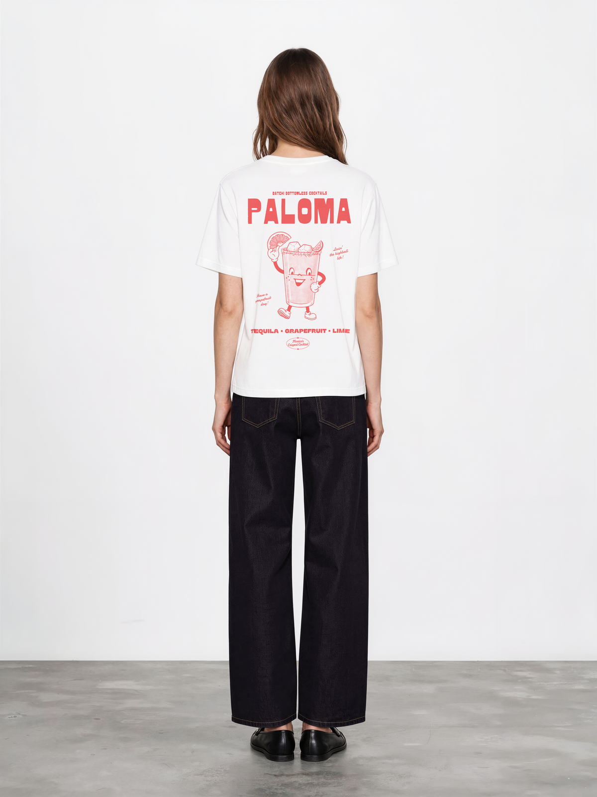 Female model wearing the Paloma white T-shirt, back view showing the large coral Paloma cocktail character and tequila, grapefruit and lime lettering.