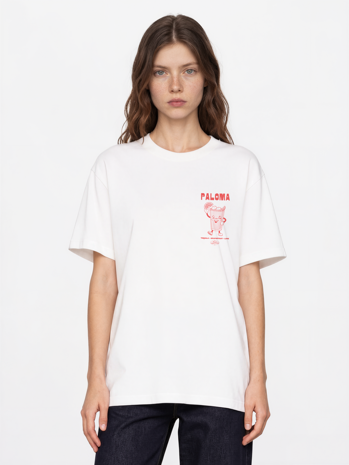 Female model wearing the Paloma white T-shirt, front view showing the coral cocktail graphic on the left chest.