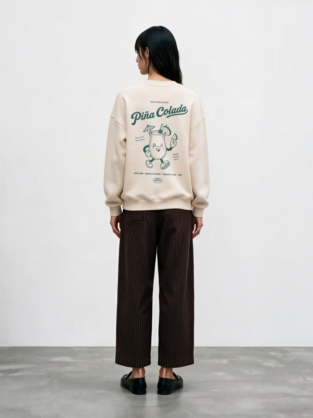 Female model wearing the Piña Colada ivory oversized sweatshirt, showing the large teal tropical cocktail character and Piña Colada lettering on the back.