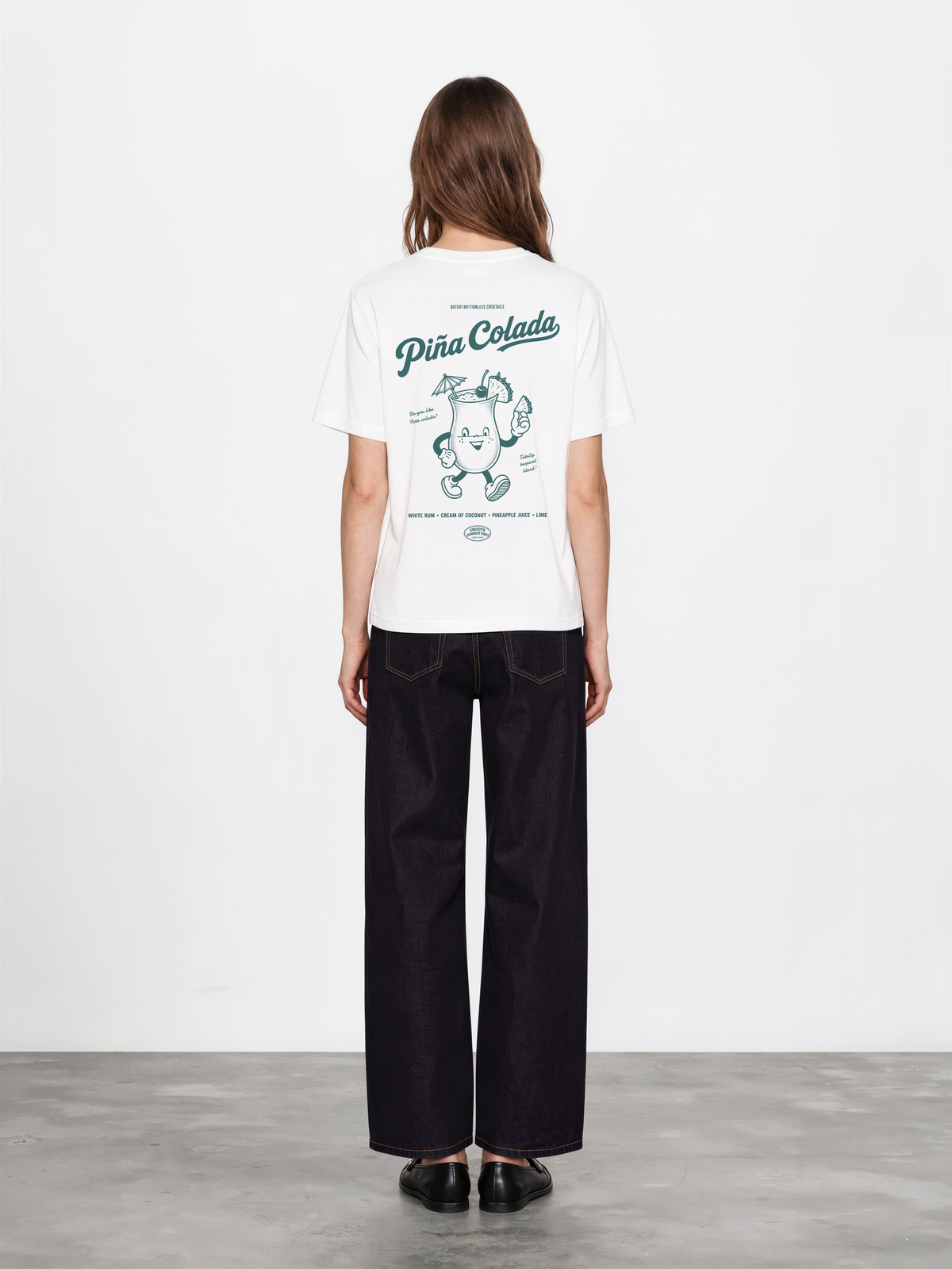 Female model wearing the Piña Colada white T-shirt, back view showing the large teal tropical cocktail graphic.