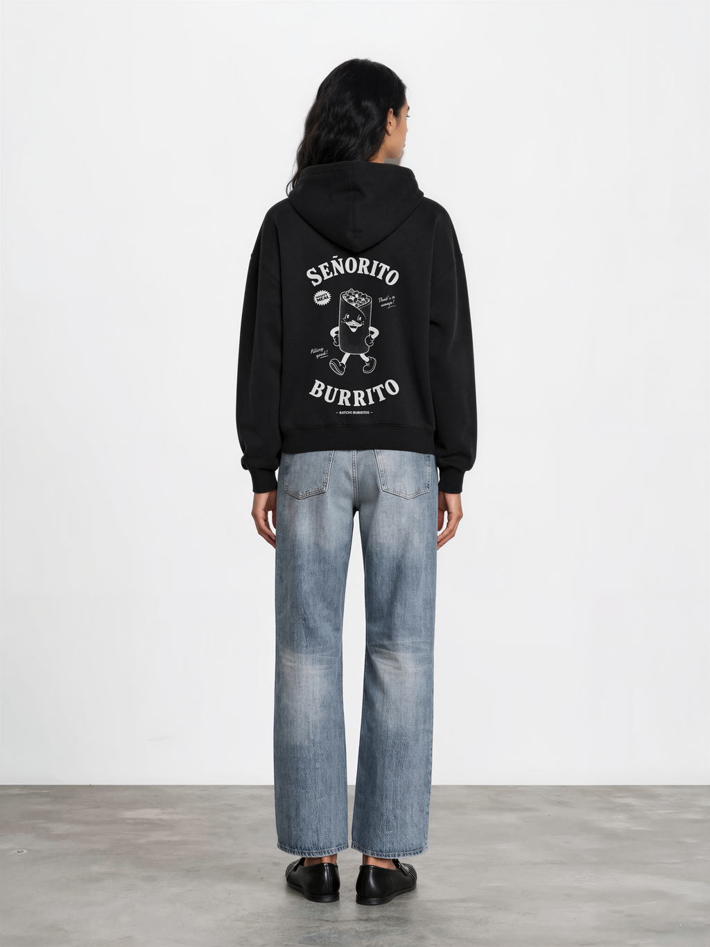 Female model wearing the Señorito Burrito black oversized hoodie, back view showing the large white burrito character and lettering beneath the hood.