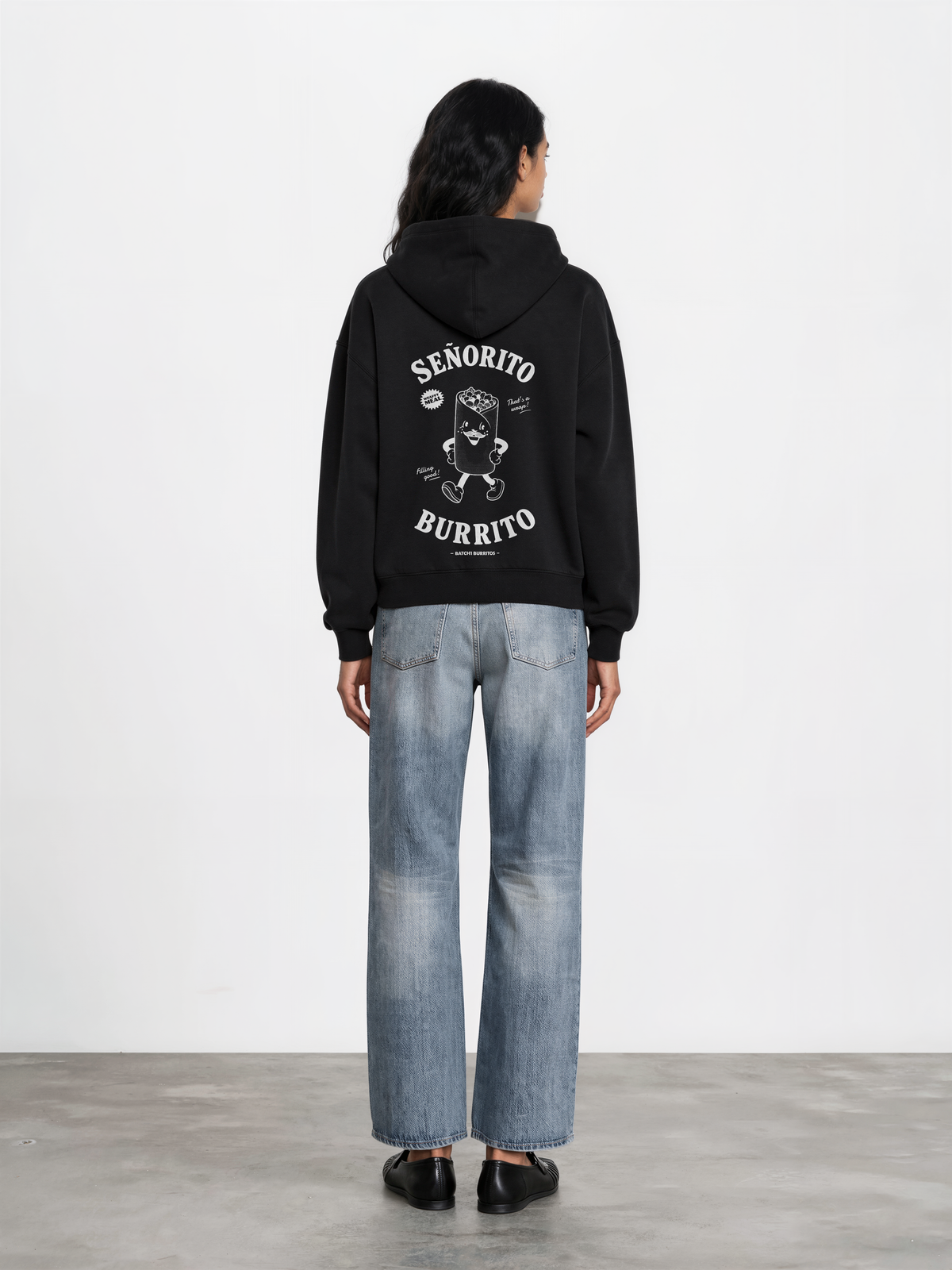 Female model wearing the Señorito Burrito black oversized hoodie, back view showing the large white burrito character and lettering beneath the hood.