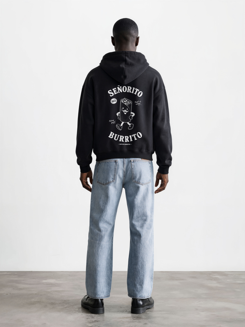 Male model wearing the Señorito Burrito black oversized hoodie, back view showing the white large burrito character and lettering.