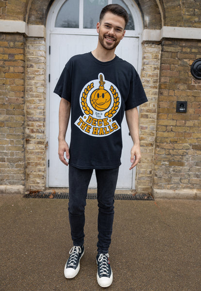 male model wears black short sleeved christmas t shirt with happy face bauble graphic and deck the halls slogan