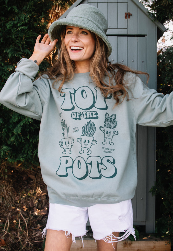 Model wears dusty green sweatshirt with Top of The Pots festival slogan and plant pot character graphic
