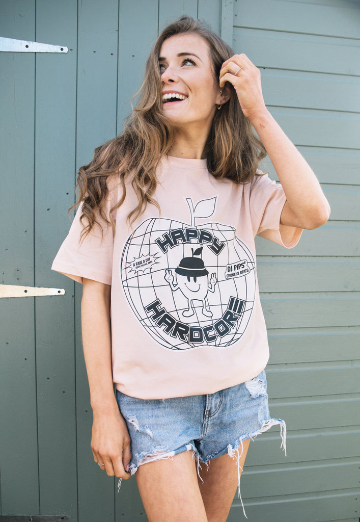 Model wears peach coloured organic t-shirt with "Happy Hardcore" slogan and apple graphics in 90s style print to front of t-shirt