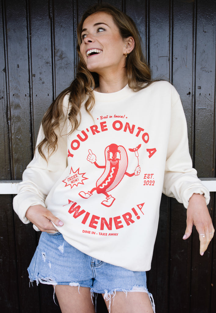 Model wears vanilla sweatshirt with You're Onto a Wiener slogan and hotdog graphic