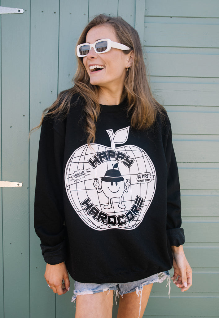 Model wears black festival sweatshirt printed with "happy hardcore" slogan and 90s style rave graphics in white